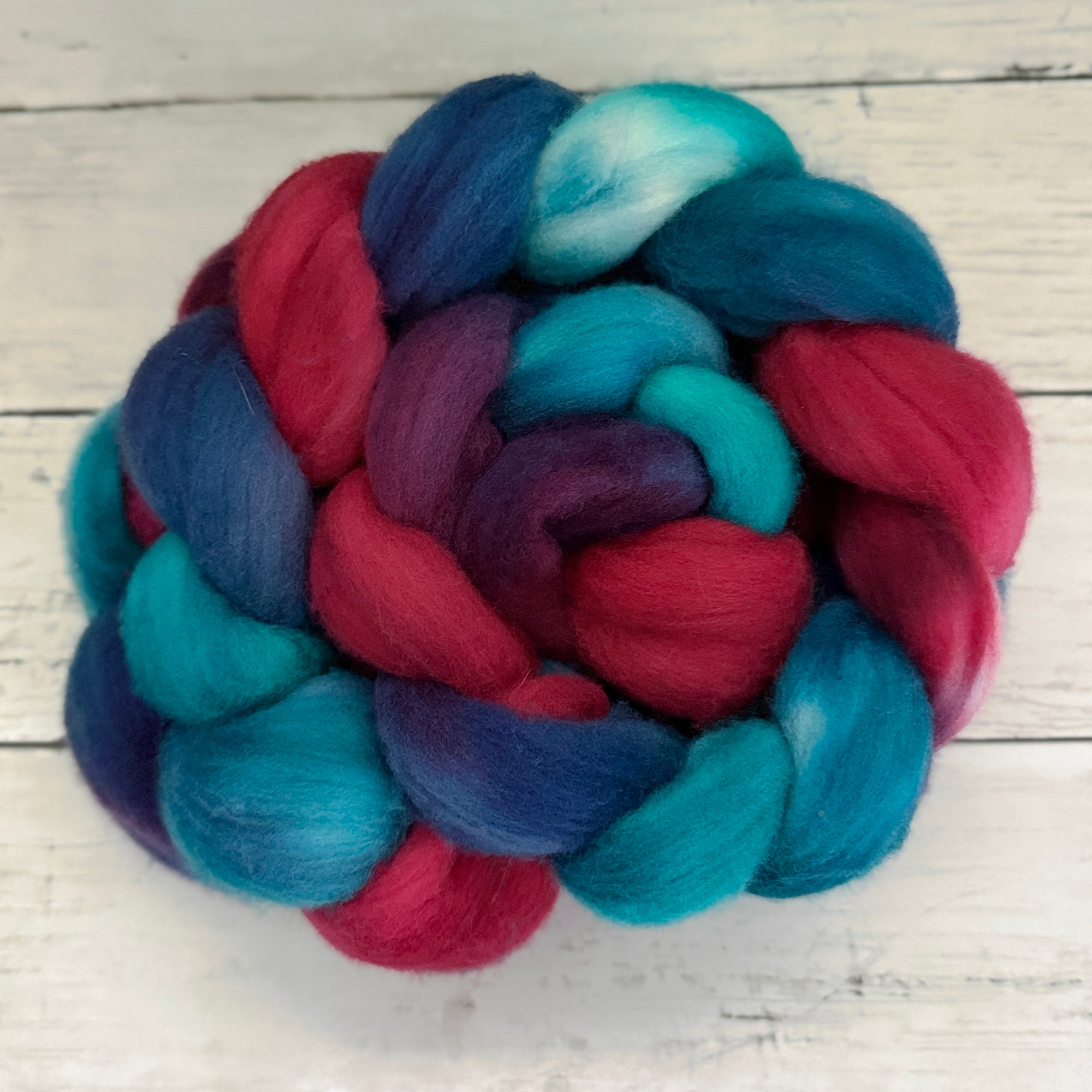A ball of variegated fiber in shades of teal, blue, purple, and magenta, braided into a decorative coil on a white wooden background.
