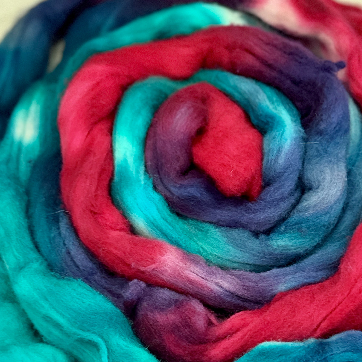 A close-up of a fluffy rosette of fiber, likely wool roving or alpaca fiber, featuring a spiral pattern of bright teal, magenta pink, deep purple, and navy blue.