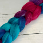 A close-up of a thick braided fiber bundle, likely wool roving or yarn, showing vibrant color transitions from bright teal and deep blue to magenta and purple, laid diagonally on a white wooden surface.