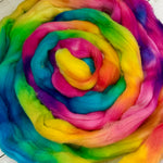 A close-up of a coiled, fluffy fiber product in a rainbow tie-dye pattern, showing vibrant bands of yellow, orange, pink, purple, blue, and green.