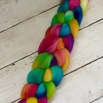 A vibrant, multicolored braided fiber bundle, likely a ponytail or braid extension, made from rainbow-colored wool roving, laid diagonally on a white wooden plank surface.