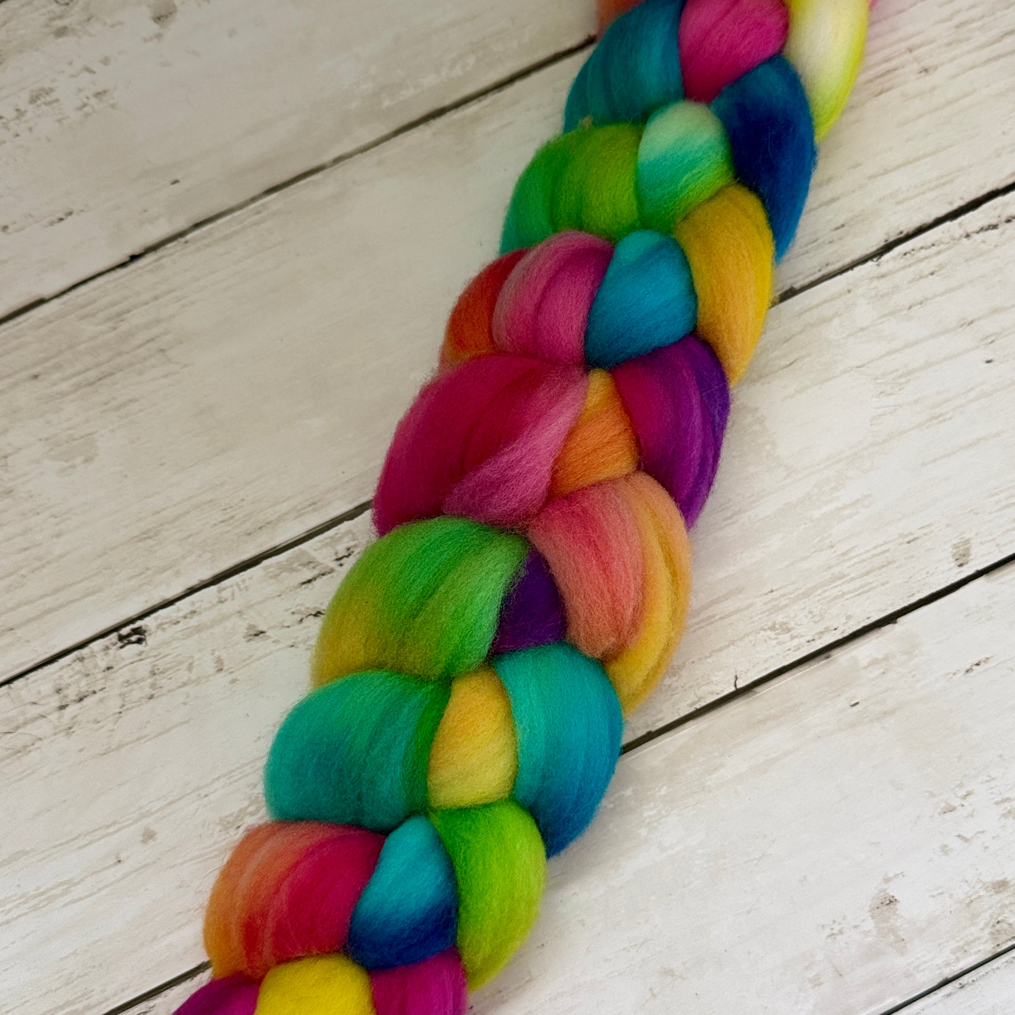 A vibrant, multicolored braided fiber bundle, likely a ponytail or braid extension, made from rainbow-colored wool roving, laid diagonally on a white wooden plank surface.