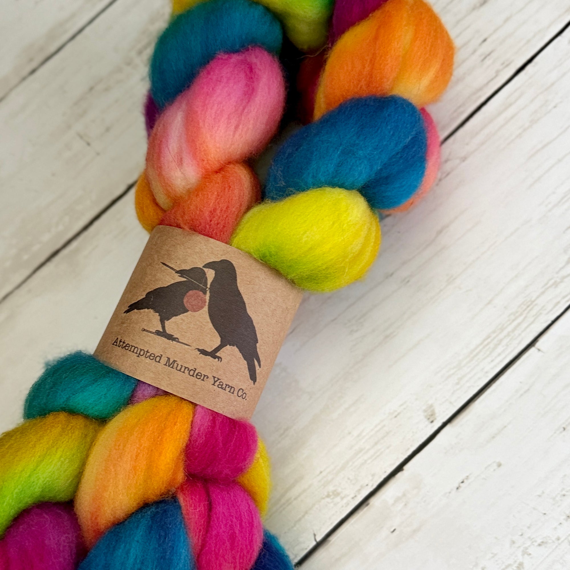 A close-up of a braided skein of rainbow-colored wool roving with a kraft paper label reading 'Attempted Murder Yarn Co.' placed on a white wood plank surface.