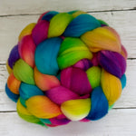 A colorful braided fiber bundle, likely wool roving or yarn, featuring vibrant rainbow shades of blue, green, yellow, pink, and purple, arranged on a white wooden surface.