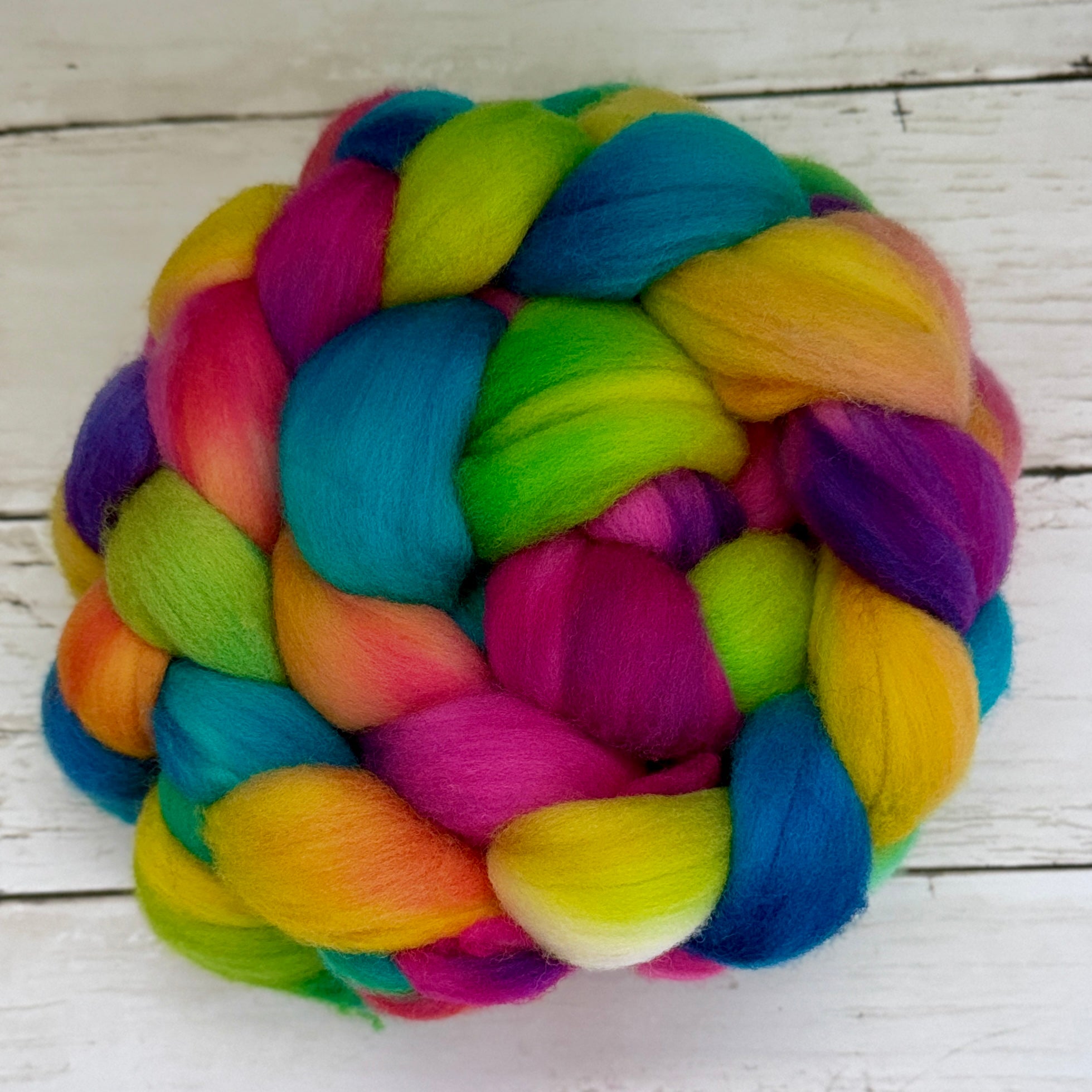 A colorful braided fiber bundle, likely wool roving or yarn, featuring vibrant rainbow shades of blue, green, yellow, pink, and purple, arranged on a white wooden surface.