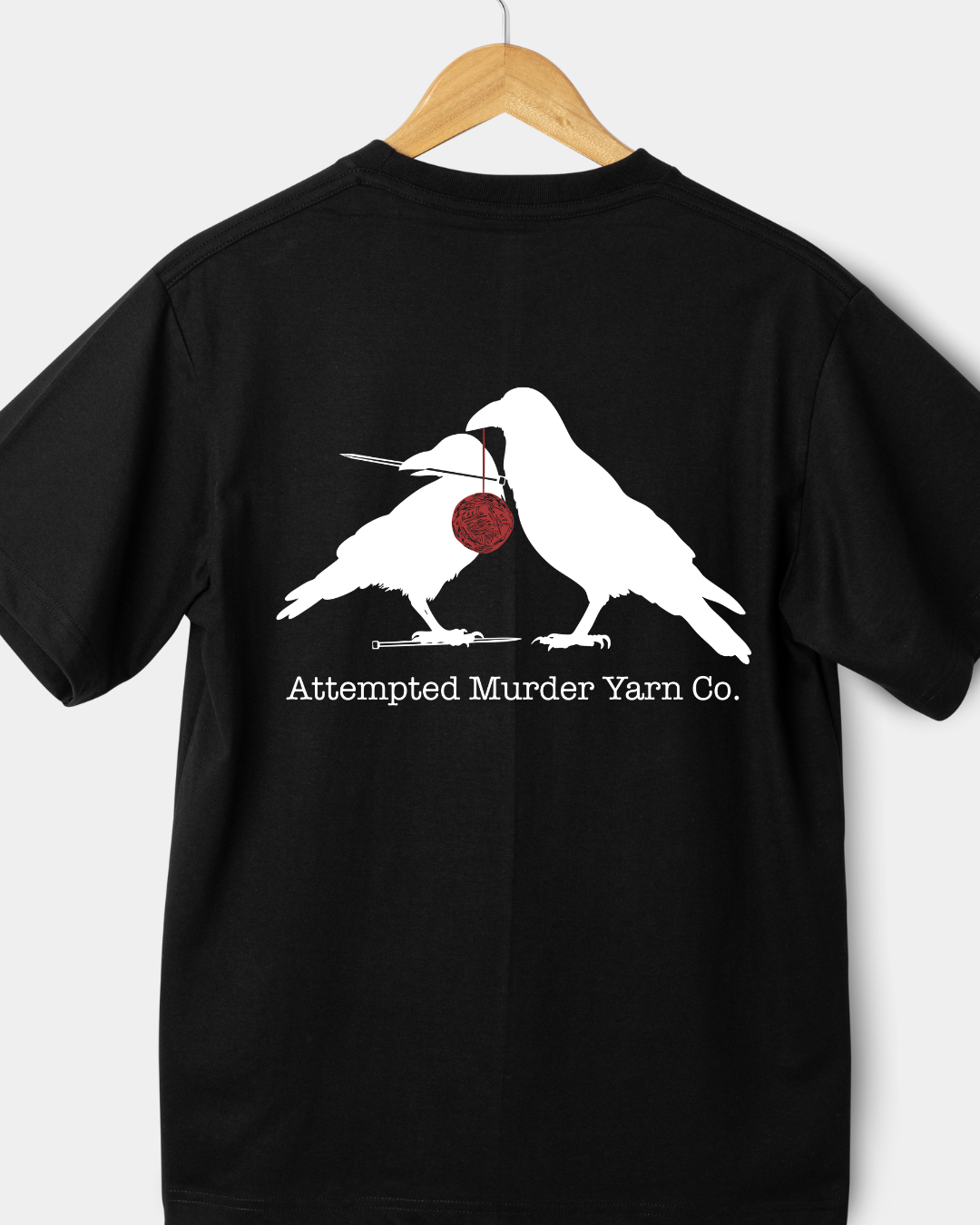 Black t-shirt with white graphic of two birds and a red yarn ball, hanging on a wooden hanger against a white background.